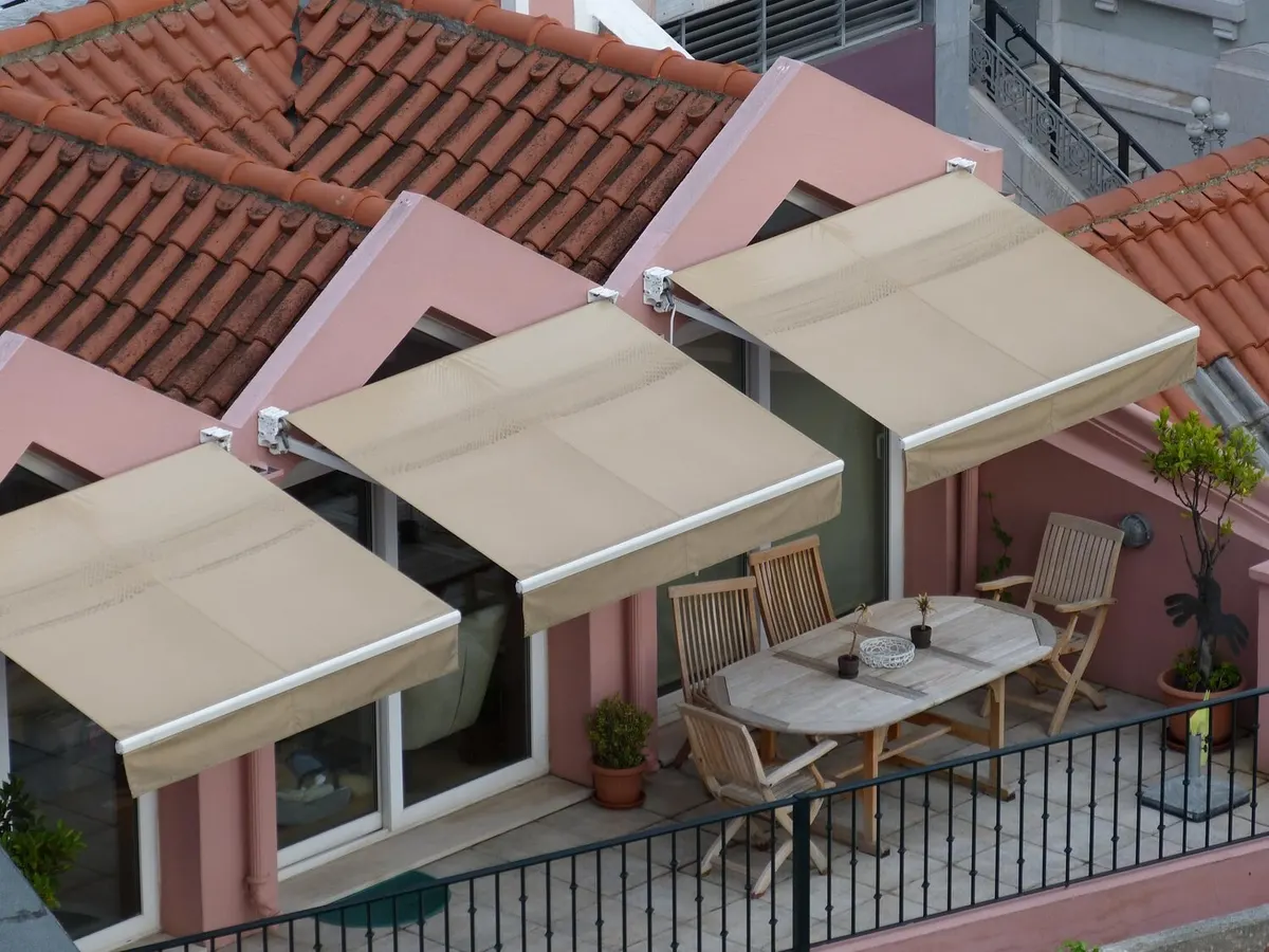 Trois stores bannes à toile beige déployés au-dessus des baies d'une terrasse, entre des toits de tuiles